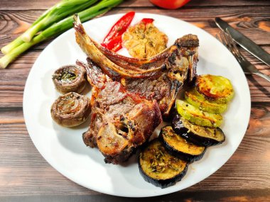 Appetizing crispy entrecote with baked eggplant, zucchini, garlic, pepper, and mushrooms on a white plate on a wooden table. A knife, fork, and green onions are nearby.