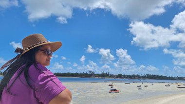 Kumsalda, kumsalda, saçları rüzgarda uçuşan siyah bir kadın. Plaj şapkası ve güneş gözlüğü takmış, doğayı düşünüyor.
