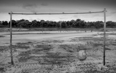 Kırsal bir futbol sahasının siyah beyaz bir fotoğrafı. Neredeyse hiç çimen ve dallardan ve bambudan yapılmış kale direkleri yok..