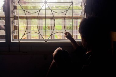 Siluet Asyalı çocuk, elleri pencere ızgaralarında oturmuş dışarıdaki ışığa bakıyor yalnızlık ve yalnızlıkla. Depresyon kavramı