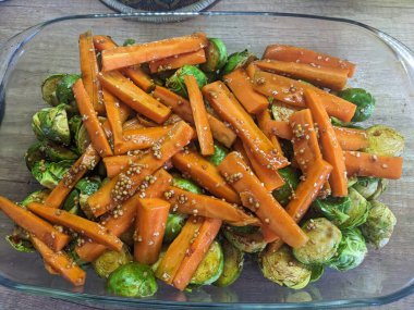 The photo displays roasted carrot pieces mixed with Brussels sprouts, seasoned with mustard and spices, all drizzled with olive oil. The vegetables are arranged in a glass baking dish, showcasing their vibrant colors and appetizing appearance.  clipart