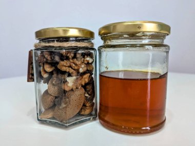 Görüntü iki cam kavanoz gösteriyor, biri altın bal ile dolu, diğeri de badem ve ceviz karışımı. Bu kavanozlar temiz bir yüzeye yerleştiriliyor. Malzemelerin doğal ve sağlıklı cazibesini vurguluyorlar.. 
