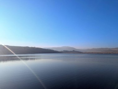 A serene view of a mountain lake captured on a sunny day, with distant peaks rising majestically along the horizon. The lake's surface mirrors the vivid blue of the sky, creating a harmonious blend of colors and light.  clipart