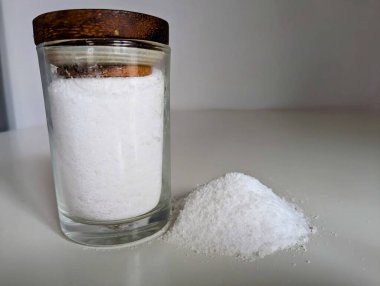 Konteynırdaki doğal tuzun yakın çekimi ve masaya konmuş küçük bir tuz yığını saf, işlenmemiş özelliklerini vurguluyor. Görüntü, doğal malzemelerin özgünlüğünü ve basitliğini vurgular.