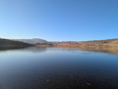 Berrak mavi bir gökyüzünün altında, suyun camsı yüzeyine mükemmel bir şekilde yansıyan bir dağ gölü. Sakin göl, uzak dağları ve kabarık bulutları yansıtıyor. 