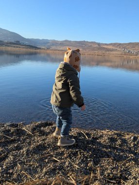 A two-year-old boy stands quietly at the edge of a pristine lake, gazing at the clear waters that reflect the sky above. Surrounded by the tranquility of nature, this tender moment captures the innocence clipart