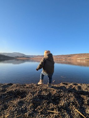 A two-year-old boy stands with quiet fascination, gazing at the still, clear waters of a pristine lake. The serene moment highlights the significance of introducing young children to the natural world, fostering curiosity and a sense of wonder.  clipart