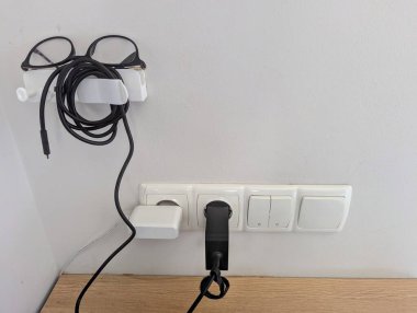 Kablolar, kulaklıklar, telefon ve şarj aksesuarları için tasarlanmış duvara monte edilmiş beyaz tutucu. Çok işlevli istasyonda düzgün depolanmış bilgisayar gözlükleri ve kulaklıklar yer alıyor.