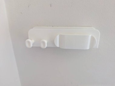 Beyaz duvara monte edilmiş kablolar düzenlemek ve akıllı telefonu güvenli bir şekilde tutmak için tasarlanmış bir tutucu. Bu pratik ve minimalist aksesuar, cihazlara kolay erişim sağlarken masaları ve başucu alanlarını dağınıklıktan uzak tutmaya yardımcı olur. 