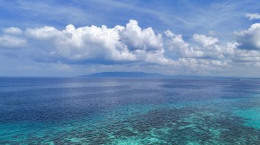 Nefes kesen tropikal deniz manzarası kristal berrak gök mavisi suları, uzak yemyeşil bir ada ve canlı mavi gökyüzüne karşı çarpıcı beyaz bulutlar. Seyahat, doğa ve okyanus için mükemmel.