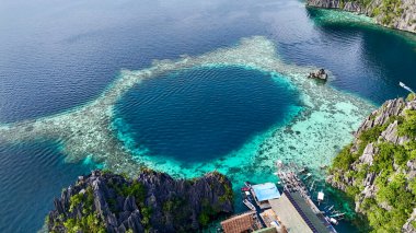 An incredible aerial view of a pristine coral lagoon with vibrant turquoise and deep blue waters. The scene is surrounded by rugged limestone cliffs covered in lush greenery, with small boats docked clipart