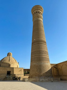 Karmaşık tuğla desenlere sahip çarpıcı tarihi bir minare, antik zamanların mimari dehasını yansıtan kubbeli bir yapının yanında duruyor. Sahne canlı bir mavi altında çekiliyor.