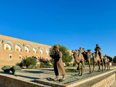 Bronze sculptures of a merchant caravan with camels in Khiva, Uzbekistan, symbolizing the historical Silk Road. The artwork is set against a traditional Uzbek architectural backdrop and a clear blue clipart