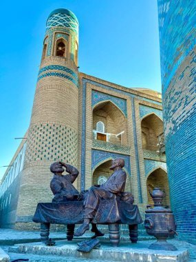 Özbekistan 'ın başkenti Khiva' da tarihi bir minarenin ve süslü mavi kiremitli duvarların güzel manzarası. Sahnede geleneksel bir platformda oturan iki adamın karmaşık İslami mimarisi ve bronz heykelleri yer alıyor.