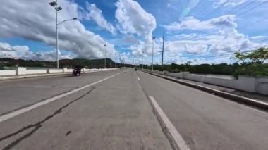 Pahalı mavi bir gökyüzünün altında, kabarık bulutlu geniş bir şehir yolu uzanıyor. Sahne yeşillik, ışık direkleri ve uzak tepelerle çevrili. Huzurlu ve huzurlu bir ortam yaratıyorlar.