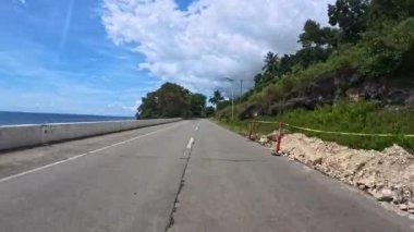 Manzaralı bir sahil yolu mavi okyanus boyunca uzanan canlı, güneşli bir gökyüzü altında, yemyeşil yapraklarla çevrili ve berrak, diğer tarafta açık deniz, seyahat ve keşif için mükemmel..