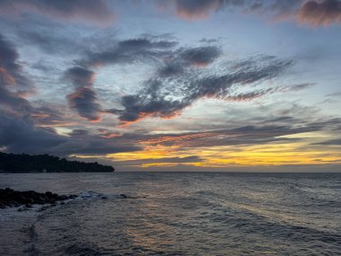 Gökyüzünü boyayan altın, turuncu ve mavi renklerin karışımıyla nefes kesen tropikal bir okyanus batımı. Dramatik bulutlar derinlik ve kontrastı eklerken, yumuşak dalgalar kayalık kıyılara dokunuyor. Siluet.