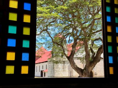 A historic stone church with a red roof is seen through a traditional window with colorful stained glass panes. A large tree with sprawling branches stands in front, casting shadows on the building clipart