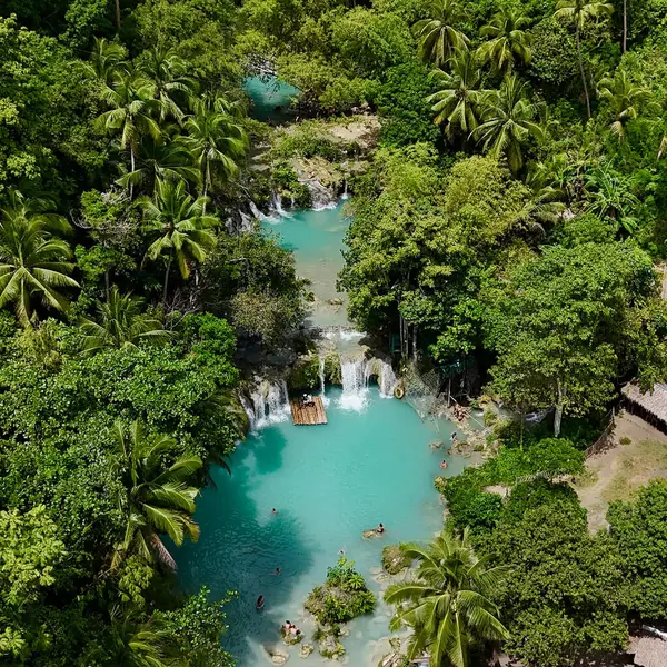 A breathtaking aerial view of cascading turquoise waterfalls surrounded by lush tropical jungle. Visitors can be seen swimming in the clear waters and floating on a bamboo raft, enjoying the natural