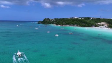 Kristal berraklığında turkuaz su ve beyaz kumlu plajları olan tropikal bir sahil şeridinin nefes kesici bir görüntüsü. Geleneksel tekneler sakin denizde süzülürken, yeşil tepeler tatil köyleri ile dolup taşıyor.