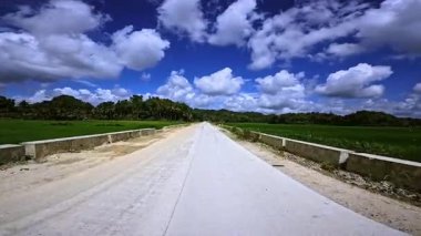 Yeşil pirinç tarlaları ve tropikal bitki örtüsüyle çevrili kırsal alanda uzun beton bir yol uzanıyor. Pofuduk beyaz bulutlu parlak mavi gökyüzü derinlik ve kontrastı