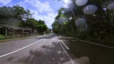 Manzaralı bir tropikal yol yemyeşil bir manzaranın içinden geçiyor, başınızın üzerinde renkli bayrak sallanıyor. Uzun palmiye ağaçları ve yoğun bitki örtüsü iyi korunmuş asfaltın her iki tarafında da bulunur.