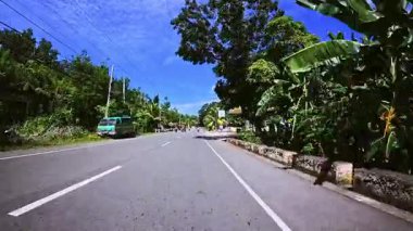 Evleri, yeşillikleri ve elektrik direkleriyle tropikal bir köyden geçen asfalt bir yol. Güneş enerjisiyle çalışan sokak lambalı küçük turuncu bir ev sağda dikilirken, gür ağaçlar ve bitki örtüsü