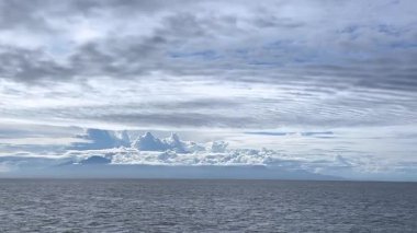 Uçsuz bucaksız okyanusun, kısmen alçak bulutlarla kaplı uzak dağlara doğru uzanan nefes kesici manzarası. Gökyüzü katmanlı bulutlarla dolu, dramatik ve sakin bir atmosfer yaratıyor.