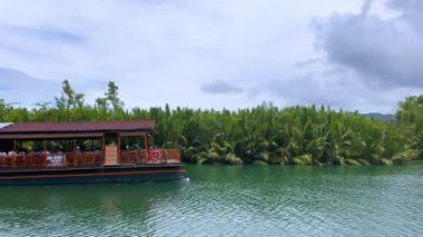 Turistleri yemyeşil alanda taşıyan yüzen bir restoran teknesi olan manzaralı bir tropikal nehir. Küçük bir motorlu tekne geziye yardım ediyor, suda hafif dalgalanmalar yaratıyor. Bulutlu gökyüzü büyütür