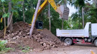 Sarı bir kazıcının tropikal ağaçlarla çevrili bir şantiyedeki beyaz bir çöp kamyonuna taş yükleyip kaldırdığı görülüyor. Olay yerinde kırsal kesimde çalışan ağır makineler görülüyor.