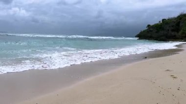 Güçlü okyanus dalgaları bulutlu bir gökyüzünün altında kumlu bir sahile çarpıyor. Sahne, dinamik su hareketi, köpüklü sörf ve dramatik kıyı şeridiyle doğanın güzelliğini yakalar. İlgili temalar için ideal