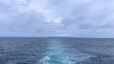 Hareketli bir teknenin arkasında dalgaların olduğu sakin bir açık deniz manzarası. Ufuk bulutlu bir gökyüzünün altında, barışçıl ve atmosferik bir deniz manzarası yaratan uzak bir adayı gözler önüne seriyor. Sahne