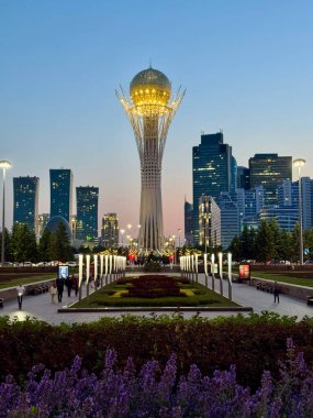Astana, Kazakistan 'daki Bayterek Kulesi' nin alacakaranlık manzarası, altın küresi açık bir akşam gökyüzüne karşı parlıyor. Ön planda ışıklı bir yaya geçidi, manzaralı çiçek yatakları ve bir...