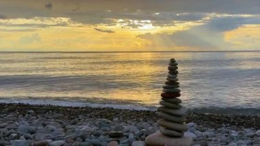 Gün batımında çakıl taşı plajında dikkatle istiflenmiş düz taş yığınının huzurlu bir sahnesi. Hafif dalgalar kıyıya vururken güneş ışınları bulutları delip sakin ve sakin bir sahil yaratıyor.