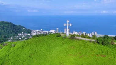 Bereketli yeşil tepeler ve mavi deniz arasında yerleşmiş canlı bir kıyı şehrini gösteren güzel bir hava görüntüsü. Yüksek binalar, yerleşim alanları ve önemli bir tepe haçı anıtı görülebilir durumda.