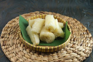 Muz yaprağı kaplı bir sepette servis edilen lezzetli buharda pişirilmiş kasava otantik ve kırsal bir mutfak deneyimi için dokunmuş bir paspasın üzerinde sunuluyor. Geleneksel gıda sunumu konseptleri ve çeşitli mutfak temaları için ideal