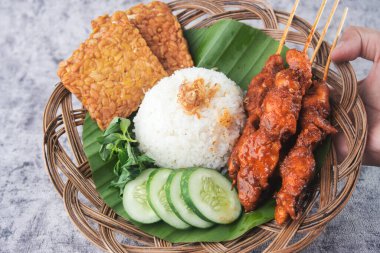 Delicious traditional cuisine served on a banana leaf platter, featuring rice, tempeh slices, skewered chicken, fresh cucumber, and leafy greens. A representation of authentic flavors and cultural culinary tastes clipart