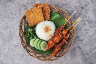 Geleneksel Endonezya yemeği tavuklu satay şiş, beyaz pirinç, tempeh ve hasır bir tabakta doğal bir yaprakla süslenmiş salatalık ile servis edilir.