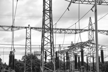 Birbirine bağlı elektrik hatları, büyük izolatörler, transformatörler ve yapısal elementler içeren yüksek voltajlı elektrik santrali, hepsi de endüstriyel altyapı ve enerji dağıtımını temsil eden bulutlu bir arkaplana karşı.