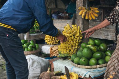 Geleneksel piyasa gösterisinde, sergilenen raflardan taze muz ve avokado seçen bireyler görülüyor.