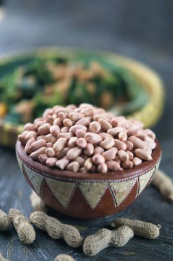 A bowl filled with shelled peanuts displayed on a textured wooden table with shells and blurred greens in the background, perfect for themes of nutrition, rustic style, or culinary ingredients clipart