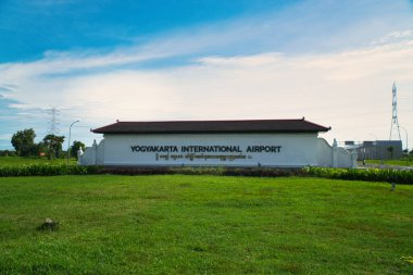 Yogyakarta Uluslararası Havalimanı giriş tabelasının manzarası, yemyeşil peyzaj ve açık mavi gökyüzü ile çevrili, seyahat ve küresel bağlantıyı simgeliyor.