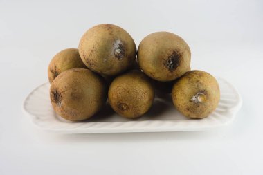 Sapodilla meyveleri arka planda beyaz bir seramik tabakta sergilenir. Tropikal yapılarını, bol miktarda sağlık faydalarını ve aşçılık ve beslenme alanlarındaki önemlerini vurguladılar.
