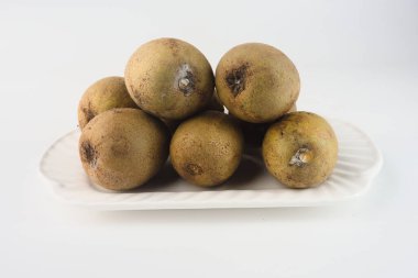 Sapodilla meyveleri arka planda beyaz bir seramik tabakta sergilenir. Tropikal yapılarını, bol miktarda sağlık faydalarını ve aşçılık ve beslenme alanlarındaki önemlerini vurguladılar.