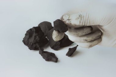 Beyaz bir yüzeye dağılmış birkaç kömür parçasıyla, karbon bazlı maddeler kullanarak kaynakları veya bilimsel deneyleri sembolize eden, bir kömür parçasını tutan eldivenli bir elin yakın çekimi.