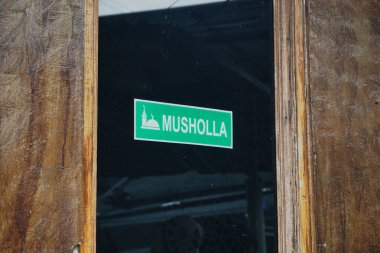 'Musholla' etiketli yeşil bir tabelanın yer aldığı, Müslümanların ibadet merkezini sembolize eden ahşap bir kapının yakın çekimi. İmaj, dinsel uygulamalara saygı ve dinginlik hissi veriyor.