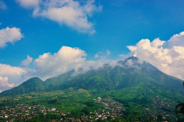 Kabarık beyaz bulutlarla çevrili yemyeşil bir Merbabu dağının nefes kesici manzarası ve açık mavi gökyüzünün arka planına kurulmuş tuhaf bir köy.
