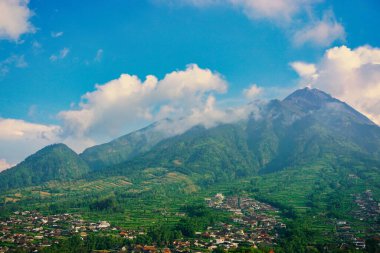 Kabarık beyaz bulutlarla çevrili yemyeşil bir Merbabu dağının nefes kesici manzarası ve açık mavi gökyüzünün arka planına kurulmuş tuhaf bir köy.