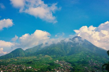 Kabarık beyaz bulutlarla çevrili yemyeşil bir Merbabu dağının nefes kesici manzarası ve açık mavi gökyüzünün arka planına kurulmuş tuhaf bir köy.