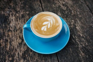 Canlı mavi bir seramik bardakta sunulan dekoratif latte, ahşap bir masa üzerinde sanatsal ve kafe estetiğini ön plana çıkarıyor.